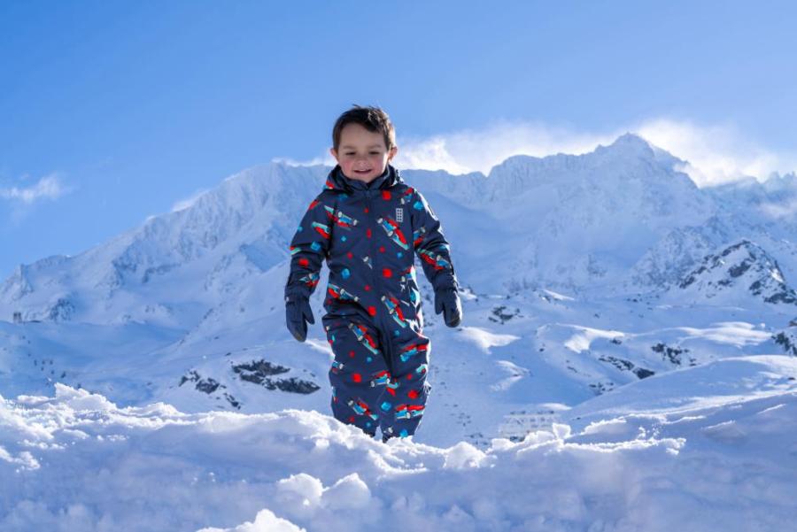 Kind in einem Schneeanzug spielt im Schnee vor verschneiten Bergen.
