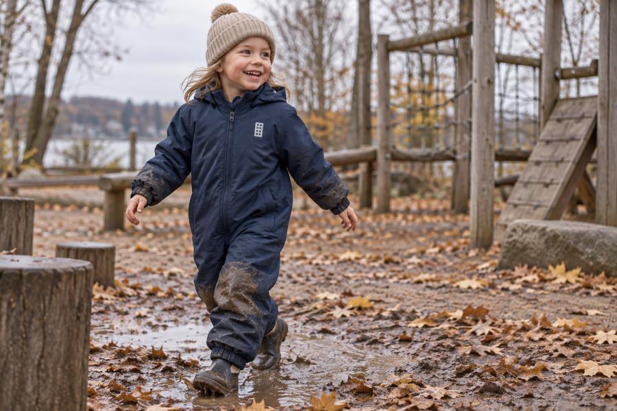 Fröhliches fünfjähriges Kind in einem schmutzigen dunkelblauen LWJORI 721 Schneeanzug auf einem Spielplatz im Spätherbst.
