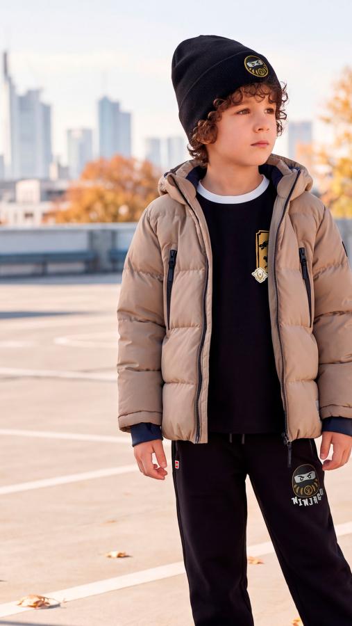 Kind in einer beigen Winterjacke und schwarzer LEGO® NINJAGO® Mütze auf einer Dachterrasse
